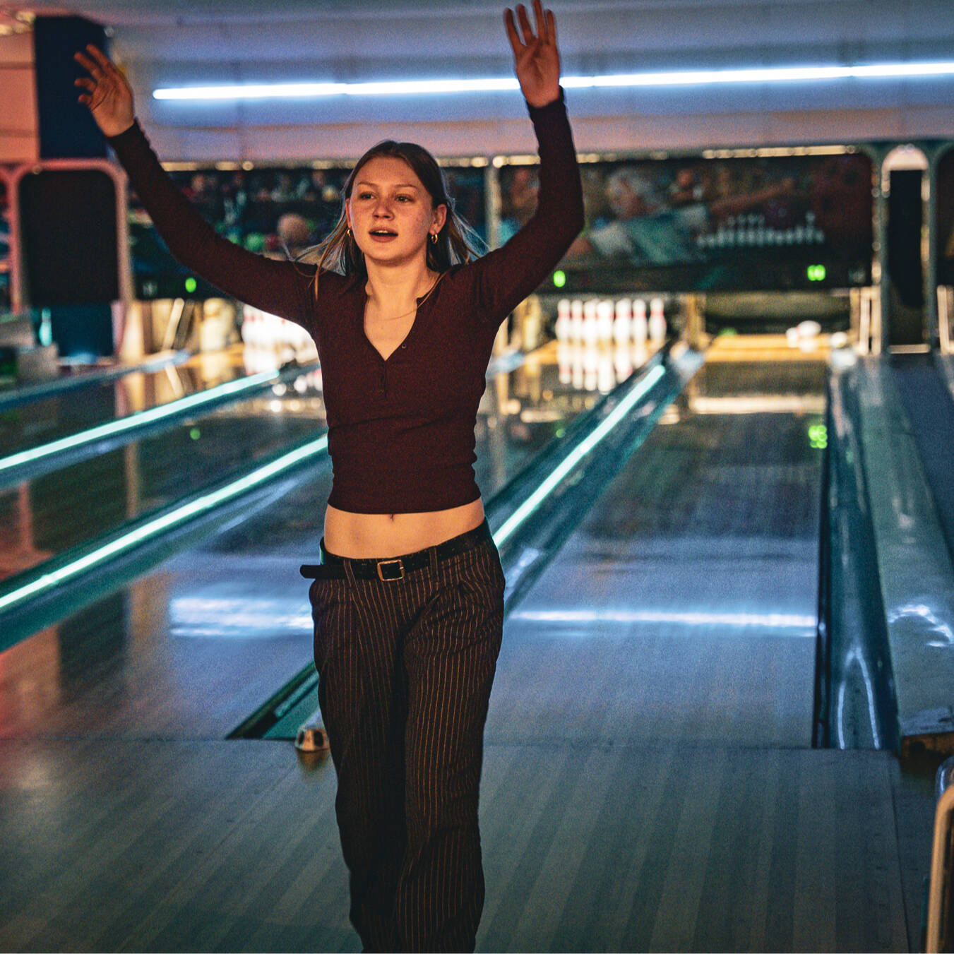 A girl celebrating with her arms in the air with a bowling lane behind her.